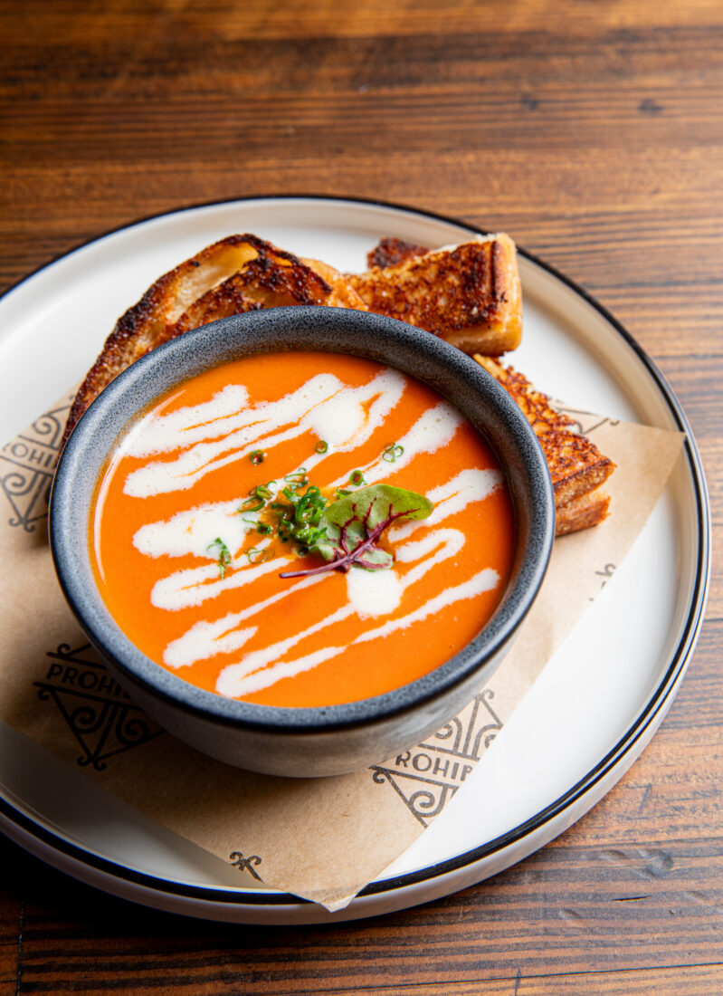 A bowl of tomato soup with a drizzle of white cream. Slices of grilled cheese are on a plate around the soup.