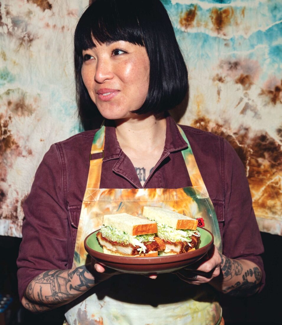 A woman in a tie dye apron holds a plate of sandwiches