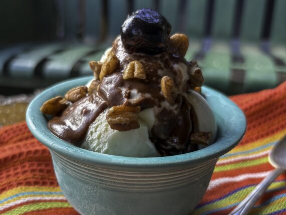 Ice cream sundae with bourbon-hot fudge, a bourbon-soaked cherry, and toasted pecans in a rustic mountain setting with a colorful striped napkin and spoon. Rusted old metal glider in background