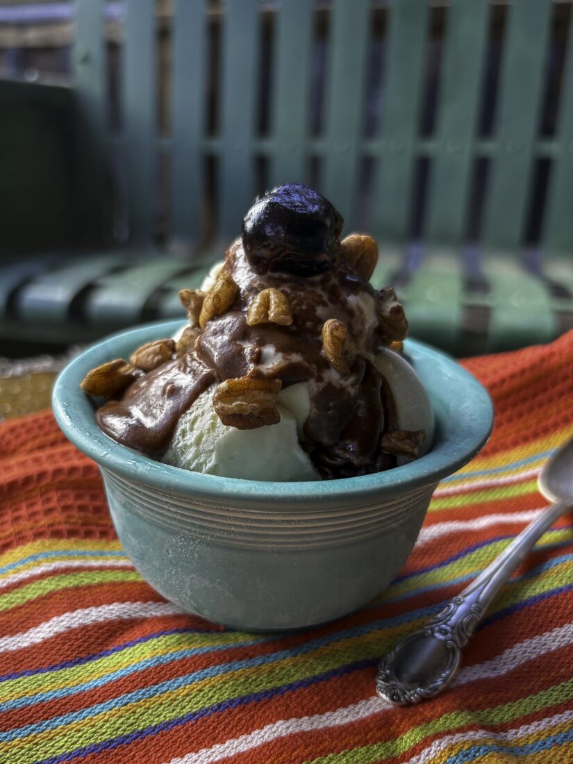 Ice cream sundae with bourbon-hot fudge, a bourbon-soaked cherry, and toasted pecans in a rustic mountain setting with a colorful striped napkin and spoon. Rusted old metal glider in background