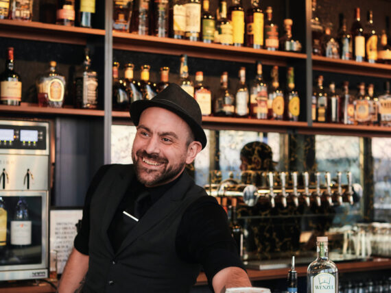 A man wearing a black vest and hat golds a whiskey sour at a black-topped bar. Walls of bottles are tucked into shelves behind him.