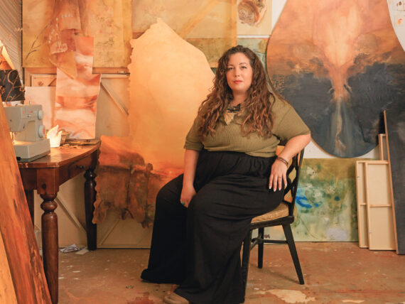 A woman sits in a chair in an art studio surrounded by earthy paintings