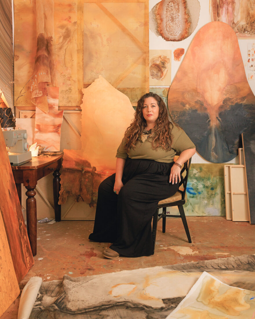 A woman sits in a chair in an art studio surrounded by earthy paintings