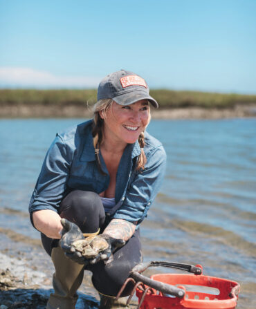 A woman harvests oysters from a marsh and smiles next to a red basket