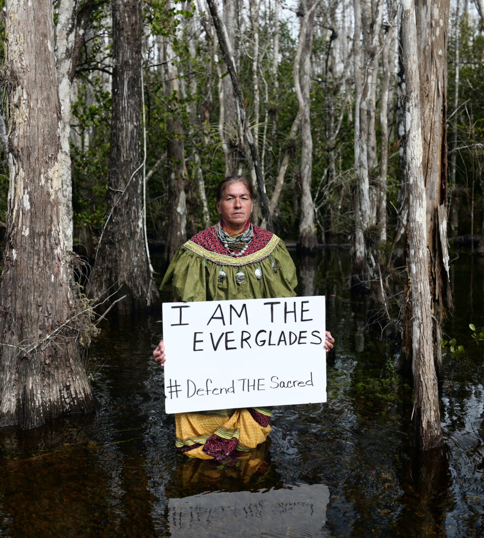 A woman stands in a swamp, in the water, with a sign