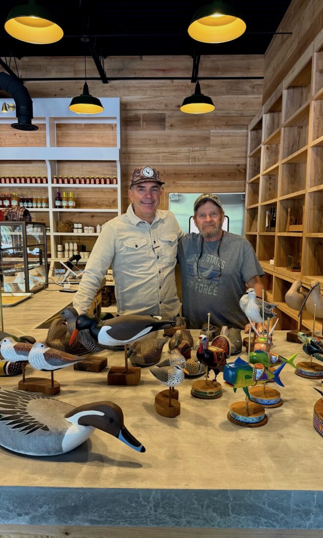 Two men stand behind a counter with decoy carvings