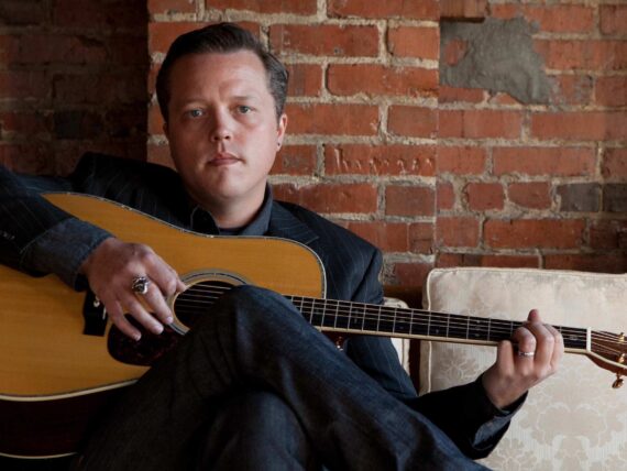A man holds a guitar and sits on a couch