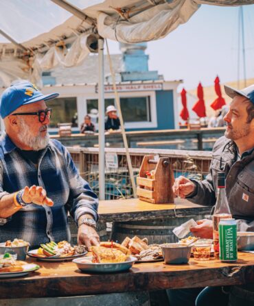 Two men sit at a table with fried seafood outside