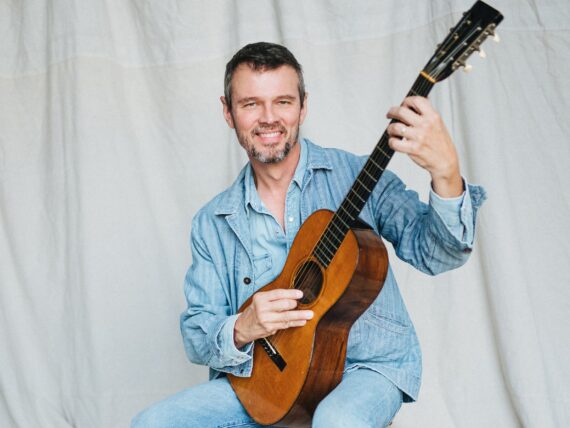 A man with brown hair, jeans, and a blue shirt holds an acoustic guitar.